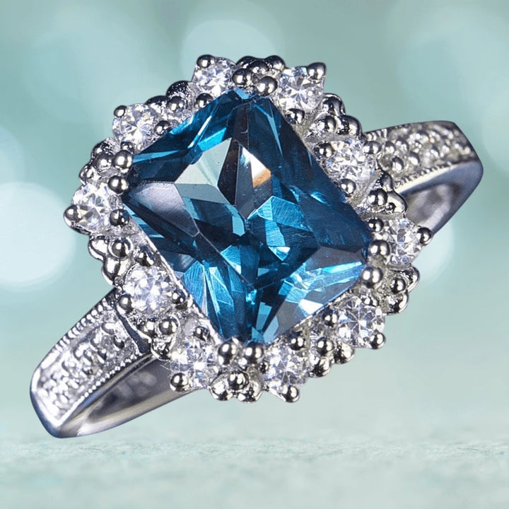 Marselles Ring featuring a large rectangular blue gemstone surrounded by smaller clear stones, set in rhodium-plated sterling silver, designed for women’s jewelry collection.