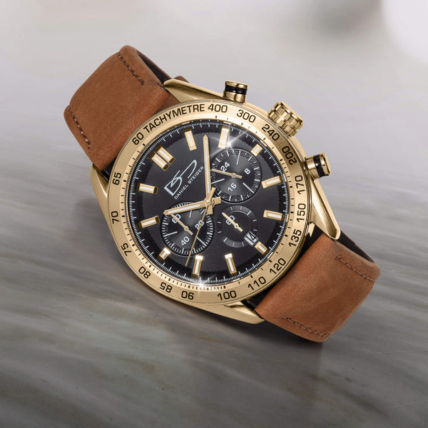 Close-up of a Daniel Steiger wristwatch with a gold-tone case, black dial featuring three subdials, tachymeter bezel, and brown leather strap, displayed on a neutral gray surface.