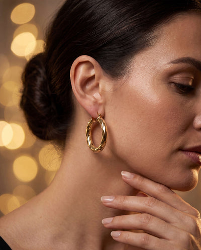Woman wearing Roma Luxe Twisted Earrings in gold-plated bronze, showcasing elegant twisted design with soft bokeh lights in background, Up To 75% Off Retail.