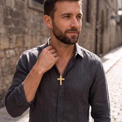 Man wearing a Phantom Gold Cross pendant on a gold chain, dressed in a dark grey shirt, shown outdoors with stone walls in the background, Up To 75% Off Retail.