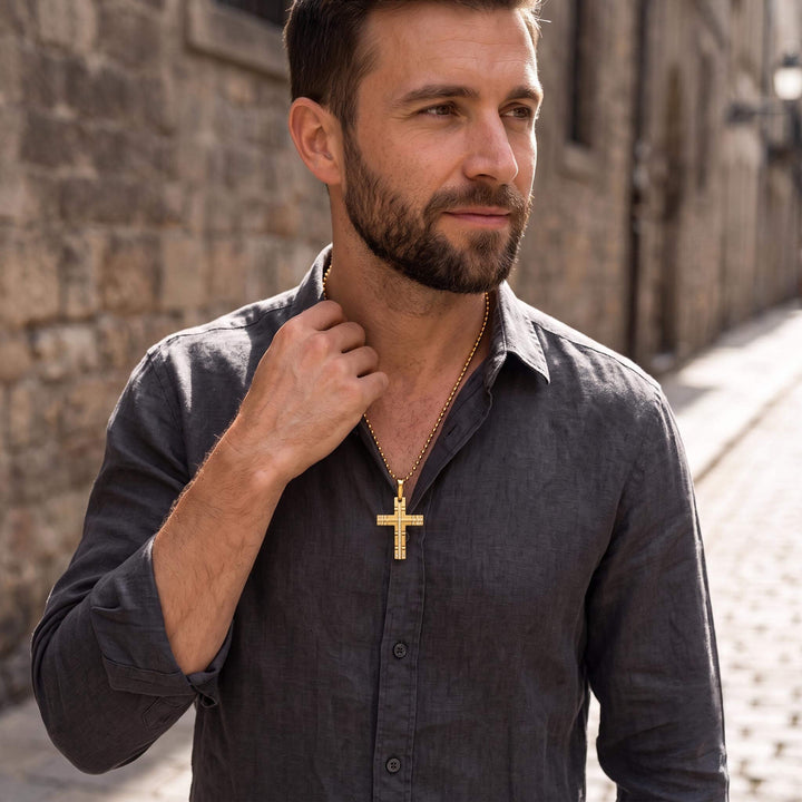 Man wearing a Phantom Gold Cross pendant on a gold chain, dressed in a dark grey shirt, shown outdoors with stone walls in the background, Up To 75% Off Retail.