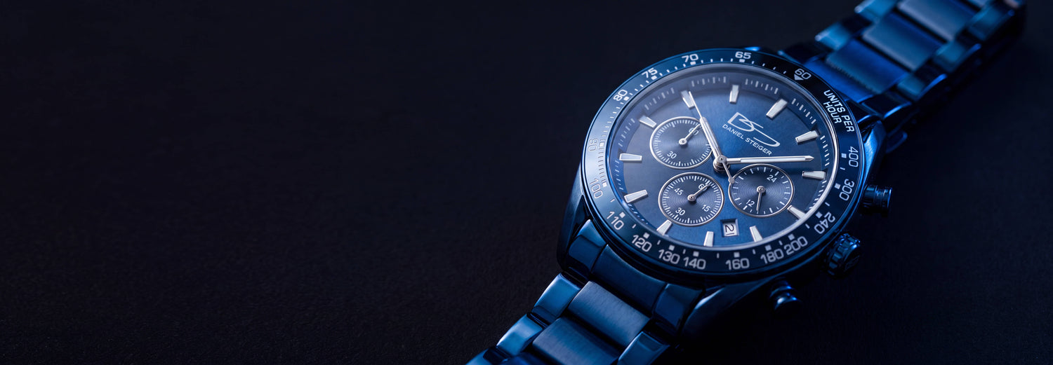 Close-up of a blue stainless steel chronograph watch with three sub-dials and a tachymeter bezel on a dark background, showcasing detailed craftsmanship and design.