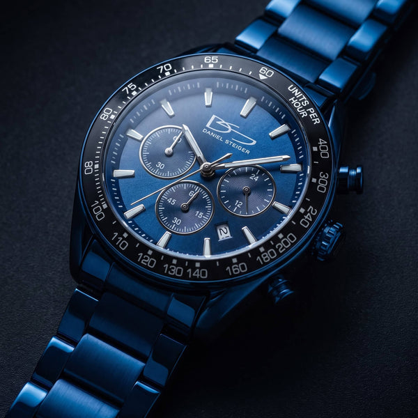 Close-up of a blue metal wristwatch with three subdials, luminous hour markers, a date window, and a tachymeter bezel on a dark background, showcasing detailed craftsmanship.