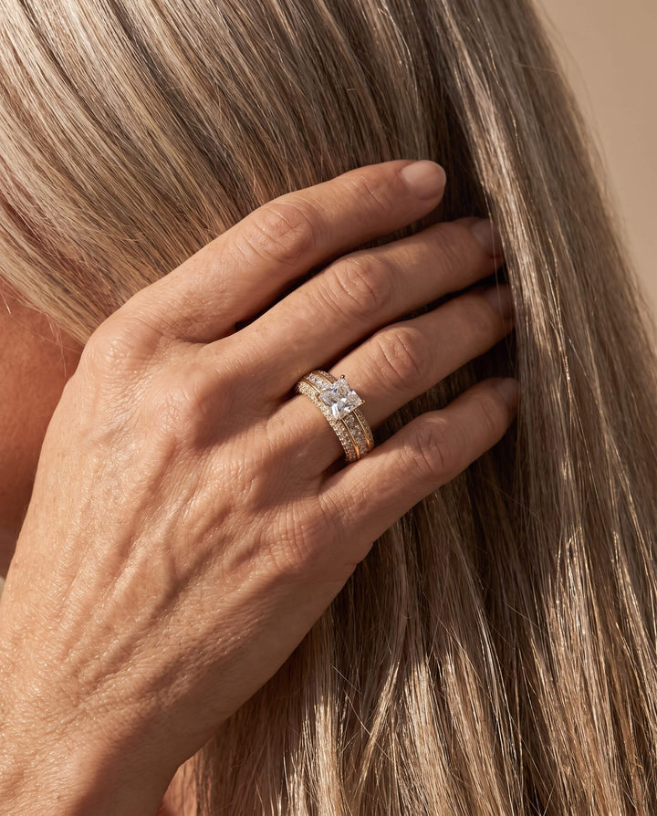Hand wearing the Charlotte Ladies Ring, featuring a sparkling square-cut center stone and multiple gold bands, highlighted against long, light brown hair, Up To 75% Off Retail.