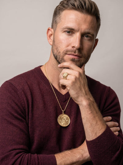 Man wearing a gold pendant necklace and matching ring with an eagle emblem, styled with a burgundy sweater, showcasing the American Glory Collection jewelry set.