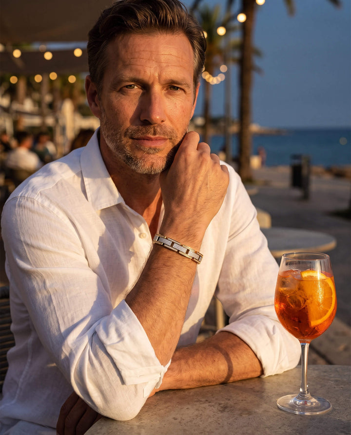 Man wearing a white shirt showcasing the Titanlink Men's Bracelet while sitting outdoors at sunset near the beach with a cocktail glass on the table, Up To 75% Off Retail.