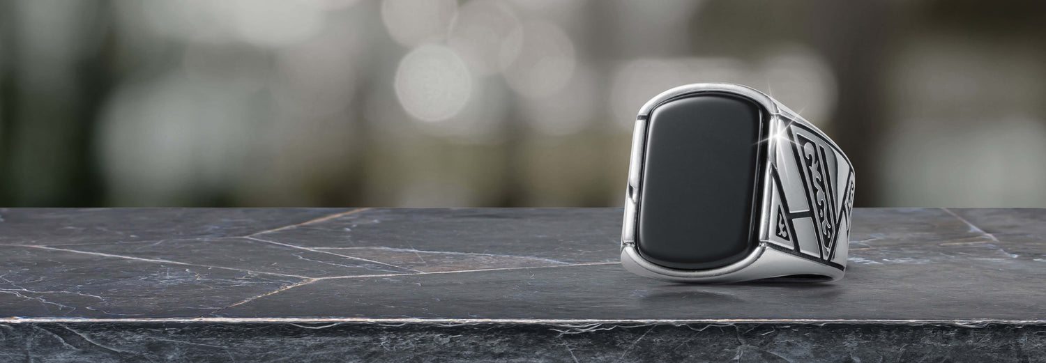 Silver signet ring with a smooth black rectangular face displayed on a dark marble surface against a blurred background, showcasing elegant jewelry design.