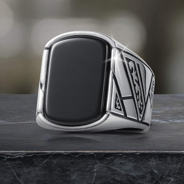 Silver signet ring with a large black rectangular stone and intricate geometric patterns on the sides, resting on a dark textured surface with a blurred background