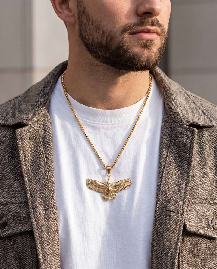 Man wearing a gold necklace with an eagle pendant, dressed in a white shirt and brown coat.