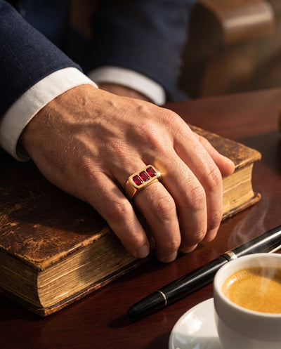 Close-up of a man's hand wearing the Sangre Men's Ring with three red stones set in a gold band, resting on a vintage book beside a pen and a cup of coffee, Up To 75% Off Retail.