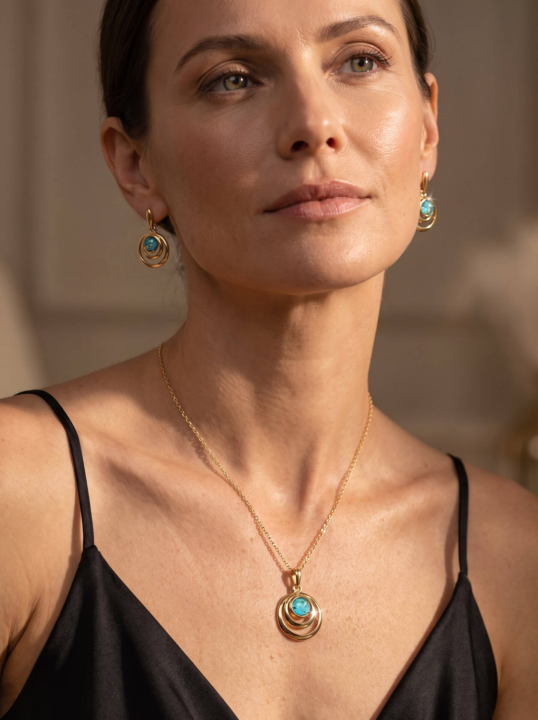 Woman wearing a gold pendant necklace and matching turquoise earrings with a black camisole in warm natural light