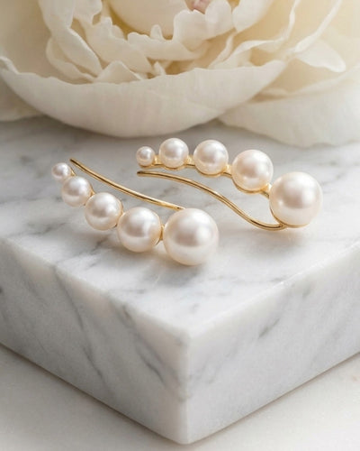 Pearl Whisper Earrings featuring a row of graduated white pearls set on elegant gold-toned curved wires, displayed on a marble surface with a soft floral background. Up To 75% Off Retail.