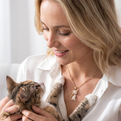 Smiling woman in a white shirt cuddles a tabby cat while wearing a delicate gold necklace with a decorative pendant. Daniel Steiger Crystelle Alley Cat Pendant