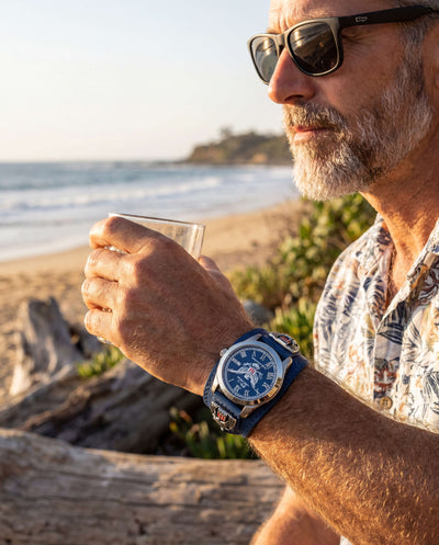 Man wearing sunglasses and a floral shirt at the beach, showcasing the Patriot 250 Years Blue Limited Edition Watch with a stainless steel case and blue dial, enjoying a drink. Up To 75% Off Retail.