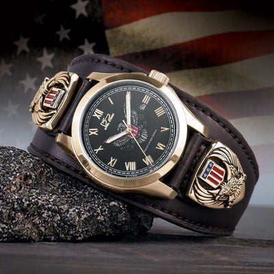 Patriot Men's Watch with gold plating, black dial, and stainless steel case, featuring a wide brown leather band decorated with American eagle and shield emblems, set against a blurred American flag background.